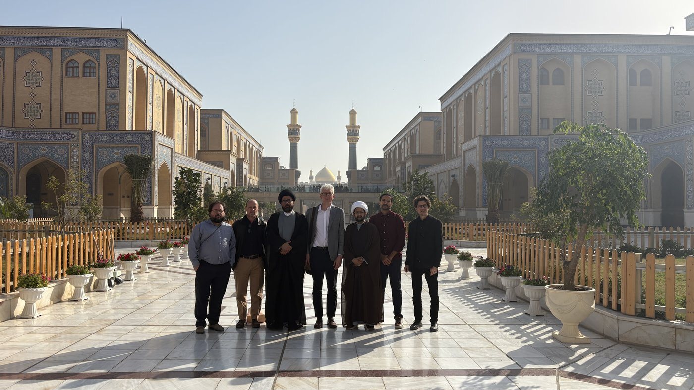 Members of the delegation, including several turbaned scholars, standing in the marble courtyard of the Shrine of Imam Ali with its golden dome and minarets behind them.